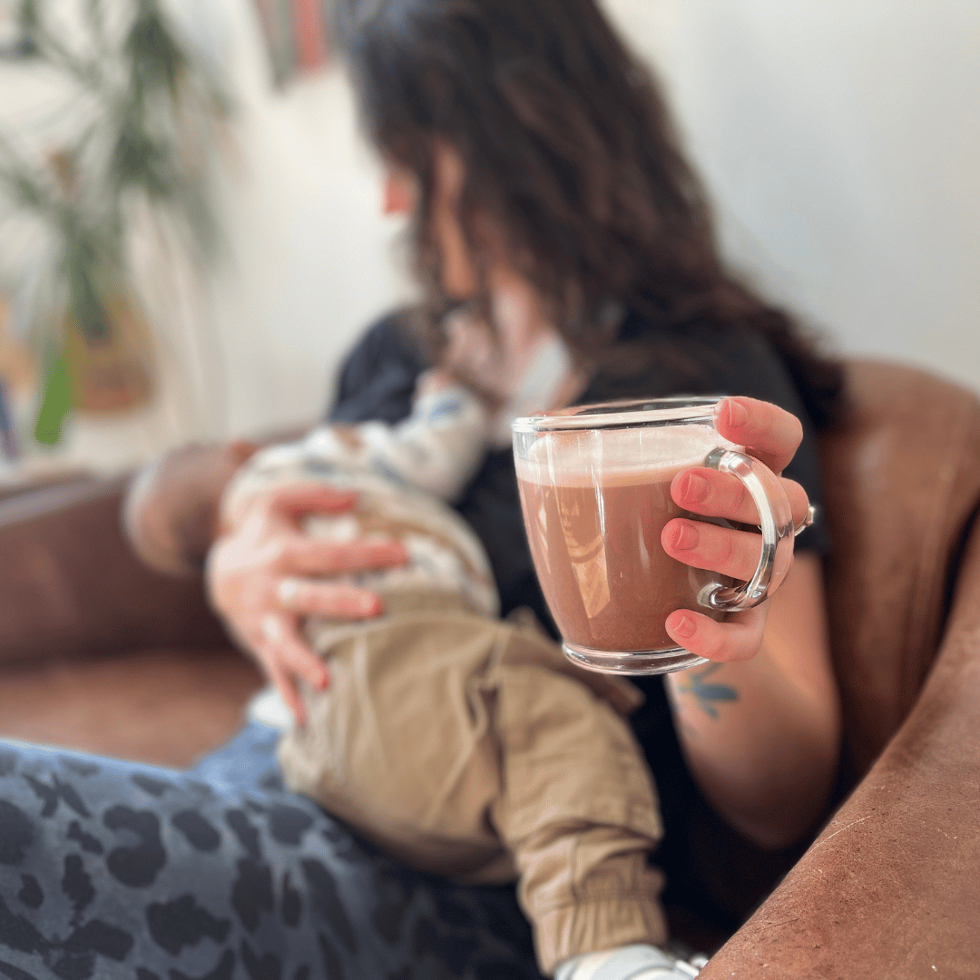 Mum sat on sofa breastfeeding her baby while holding out a cup of lactation hot chocolate to the camera