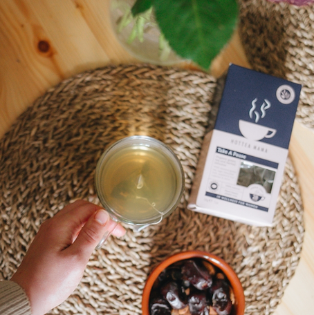 Zoomed in shot of hand holding glass tea cup of green tea, by a pack of HotTea Mama Take A Pause menopause tea, and a bowl of dates