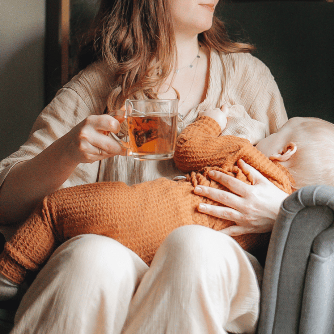 Image of a lady sitting breastfeeding her baby with a cup of HotTea Mama Milk's Up tea in her hand