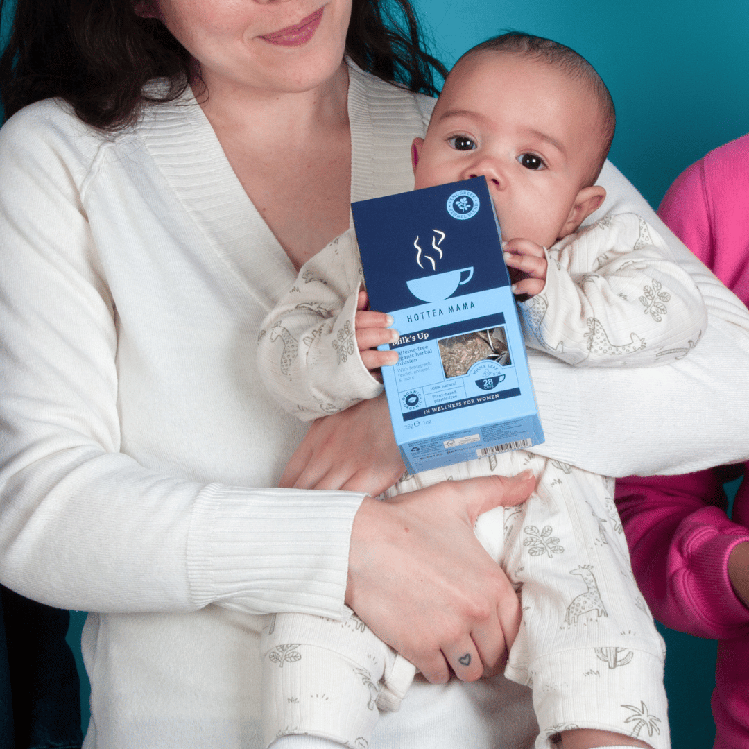 Mom in white top holding a baby who is chewing on a pack of HotTea Mama's Milk's Up breastfeeding tea