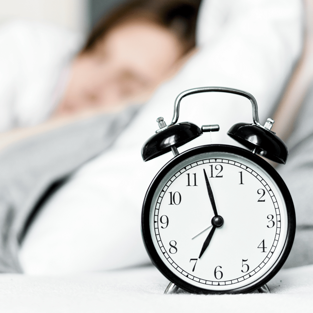 Image of a alarm clock in the forefront with a lady in the background blurred sleeping