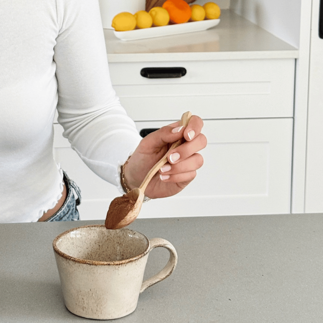 Person stirring Collagen Calm Hot Chocolate powder in a mug with a wooden spoon in a kitchen setting