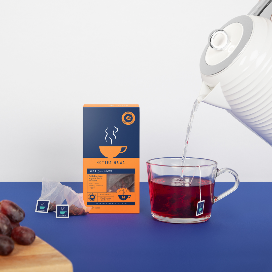Get Up & Glow organic fruit tea pack sat on blue background, with kettle filling glass cup of fruit tea, and grapes sat on chopping board near by