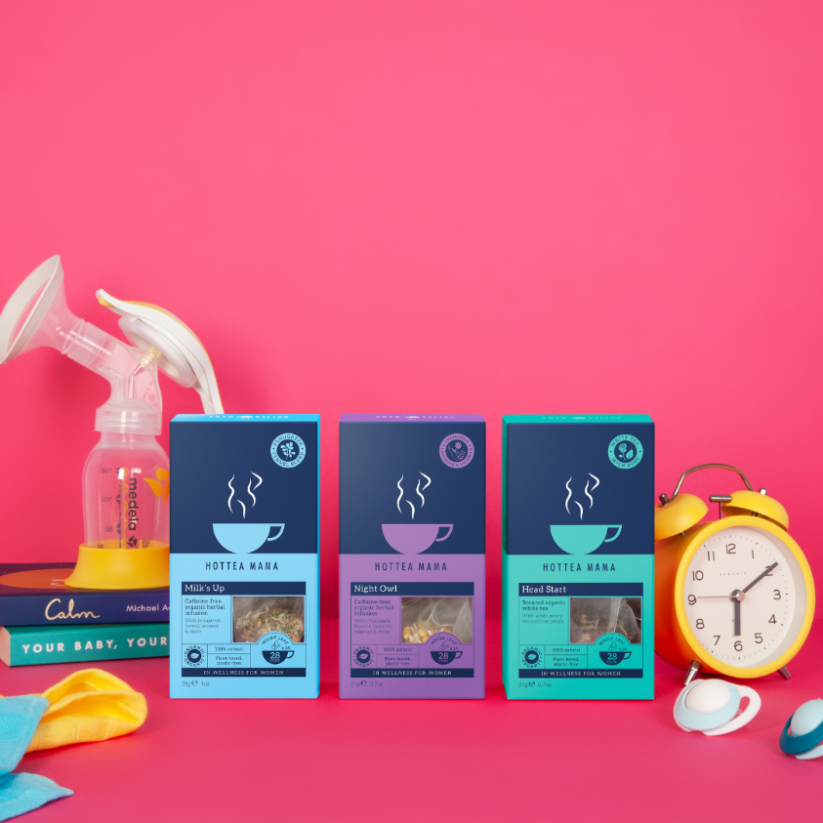 Three packs of HotTea Mama tea against a pink background, with a clock on one side and a breast pump and parenting books on the other. The teas are a blue pack of Organic Milks Up breastfeeding tea, a purple pack of Organic Night Owl tea and a turquoise pack of Head Start white tea