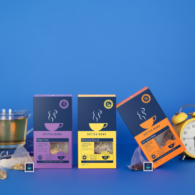 Three new mum support teas on a blue background - a brewed cup of organic Night Owl tea on top of a book about calm, Morning Rescue tea pack in middle of the shot and a pack of Get Up And Glow fruit tea on an angle next to an alarm clock