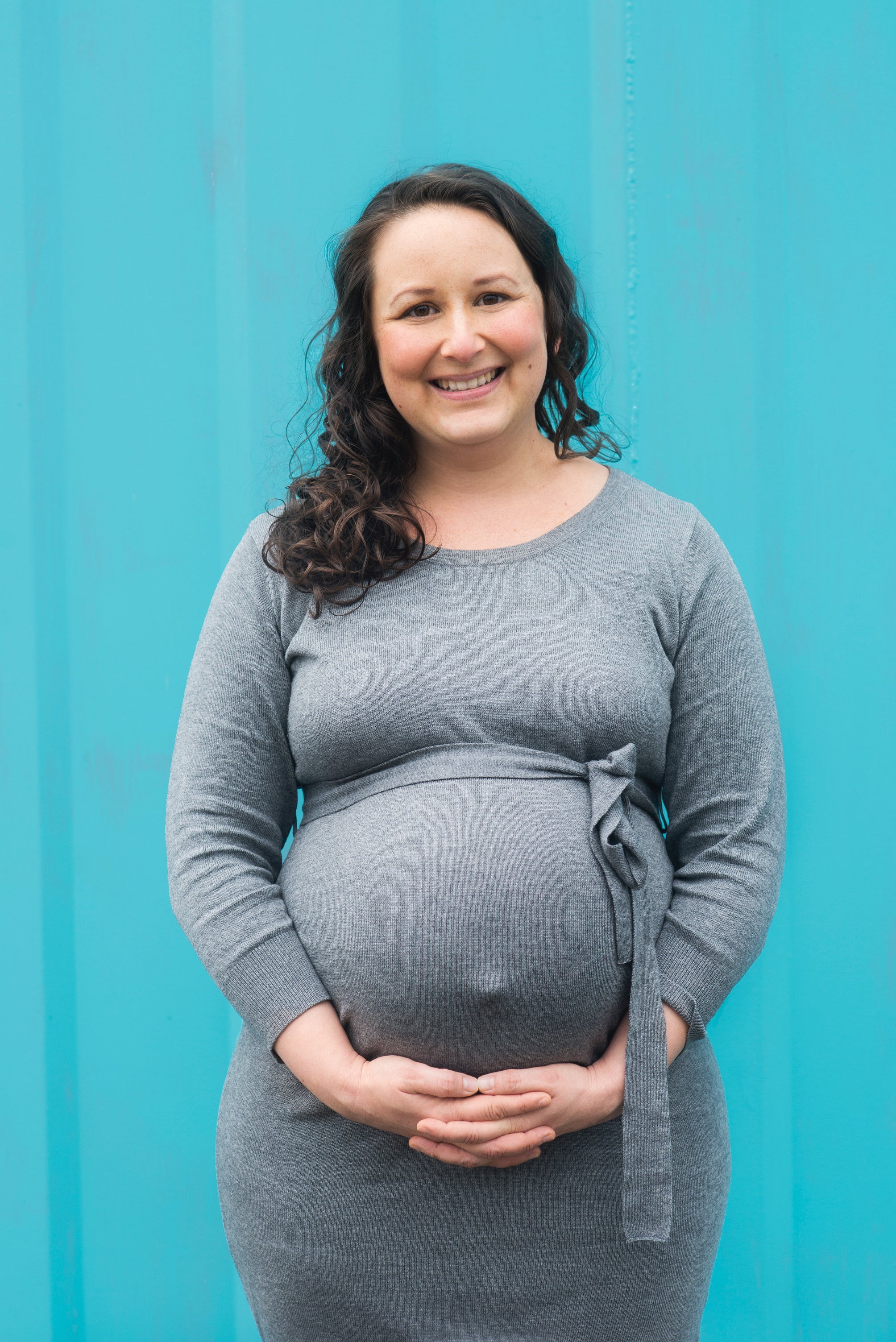 pregnant lady in grey dress, with hands under her bump, standing against a turqouise wall