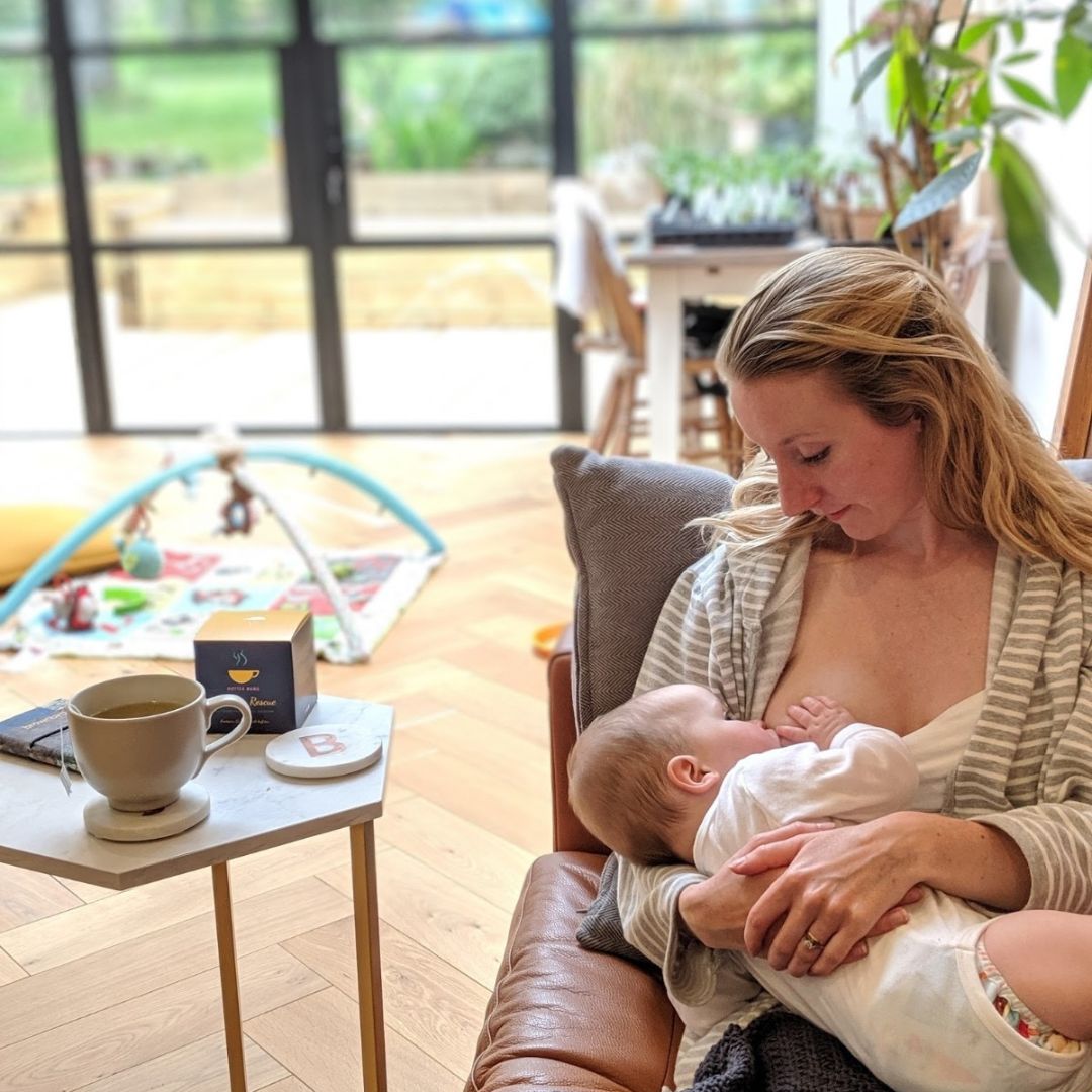 Mum in sunny room, on sofa breastfeeding her baby with table next to her with tea on it.