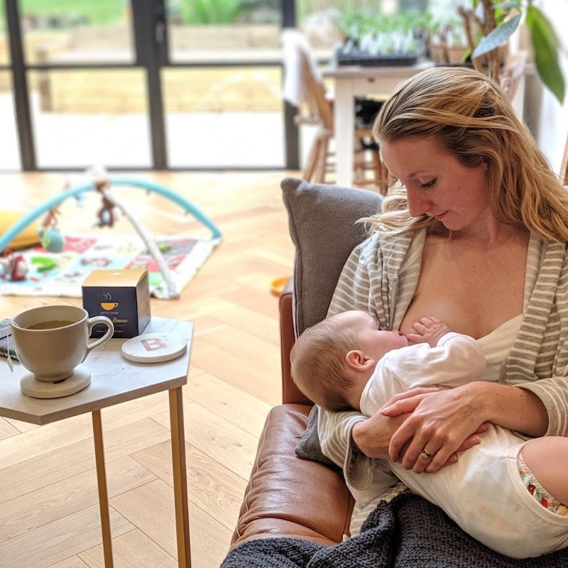Lady lying on sofa breastfeeding her baby with HotTea Mama lactation support tea on table next to her