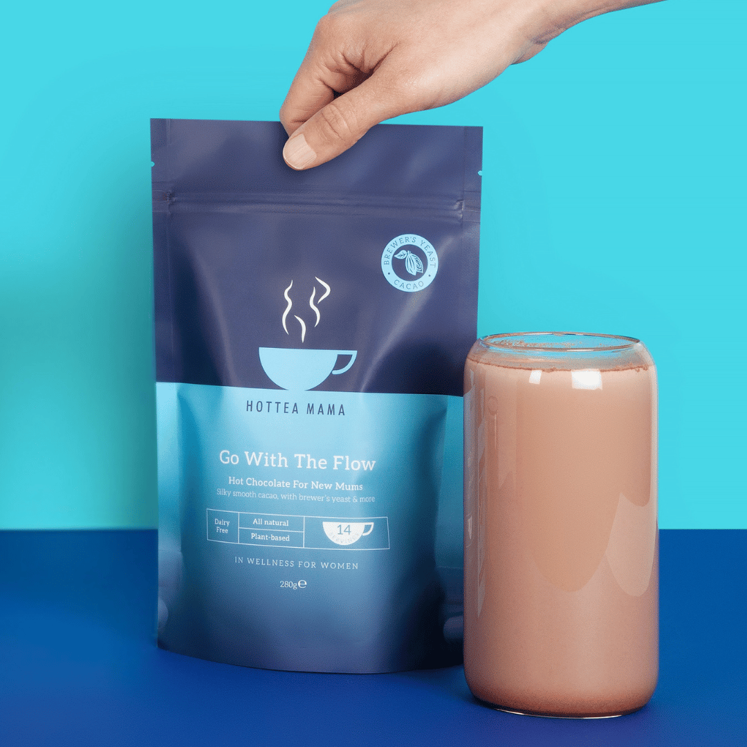 Hottea Mama Go With The Flow breastfeeding hot chocolate pack next to a glass of chocolate milk shake, on a blue background with a hand grabbing the zip lock bag