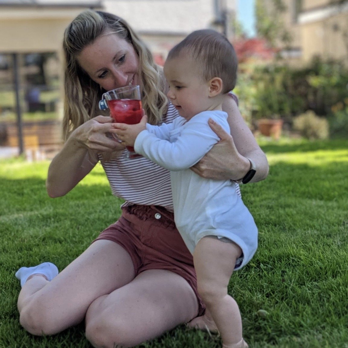 Mum in sunny garden sat down with her toddler, offering them a glass of iced tea