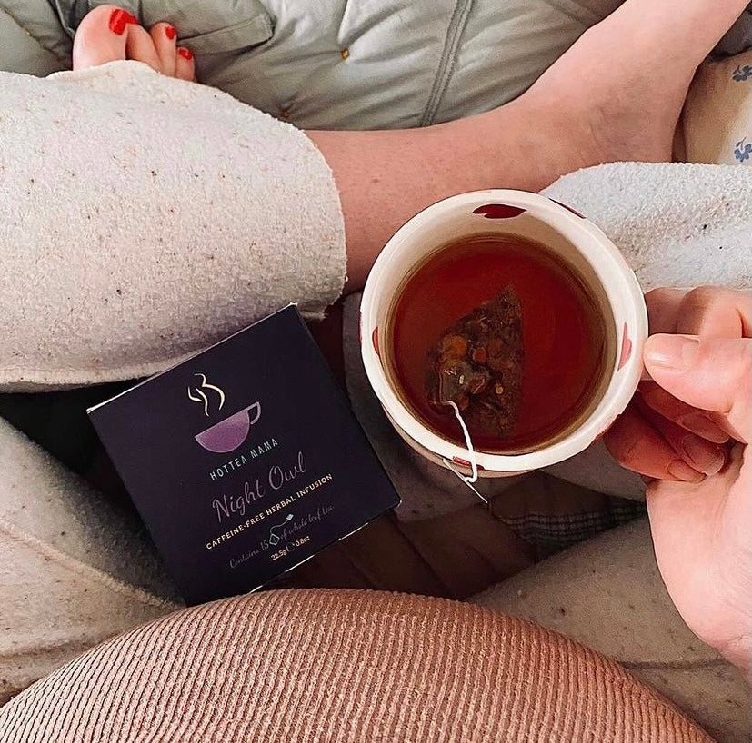Cup of night owl chamomile tea held by a lady with legs crossed, next to pregnant belly