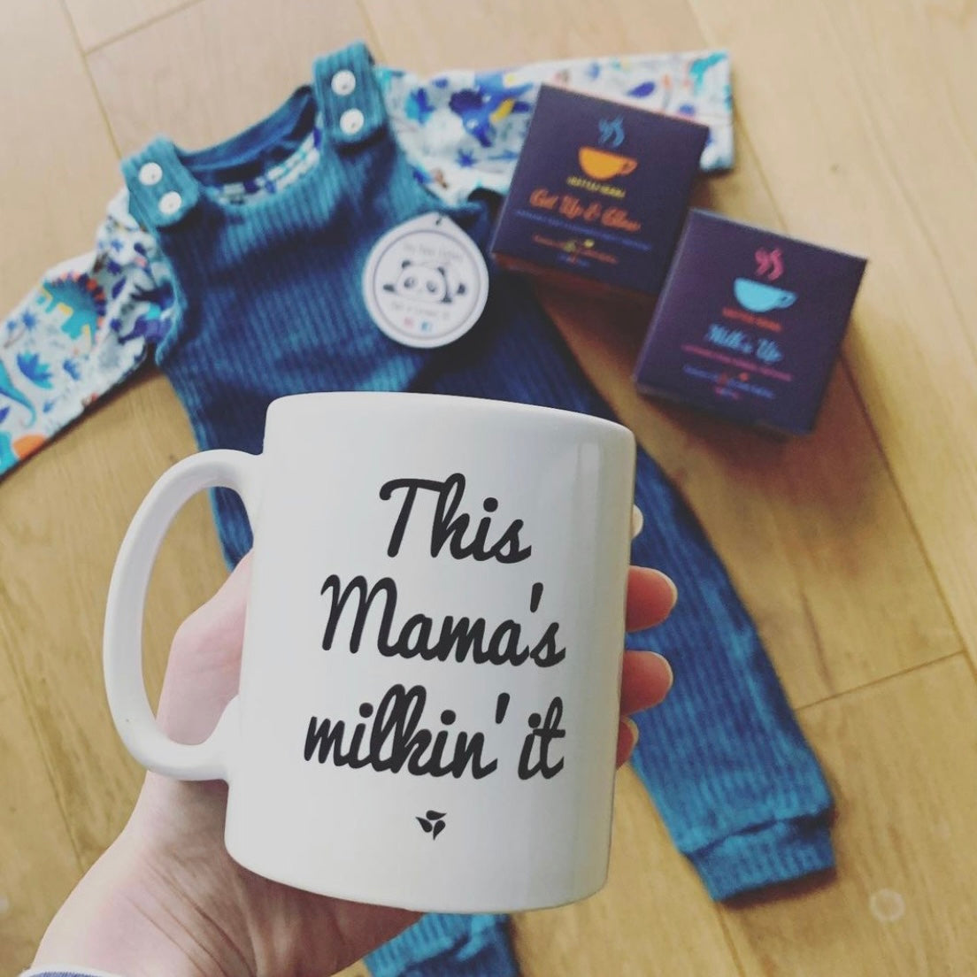 Hand holding a mum that says 'This Mama's milkin' it' in front of baby clothes and 2 packs of tea