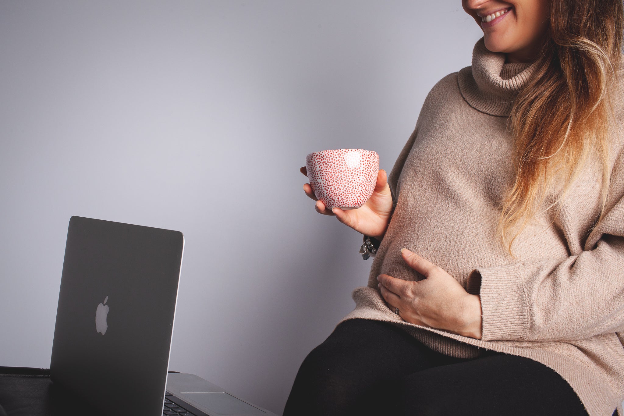 Pregnant lady sat holding cup of tea next to laptop