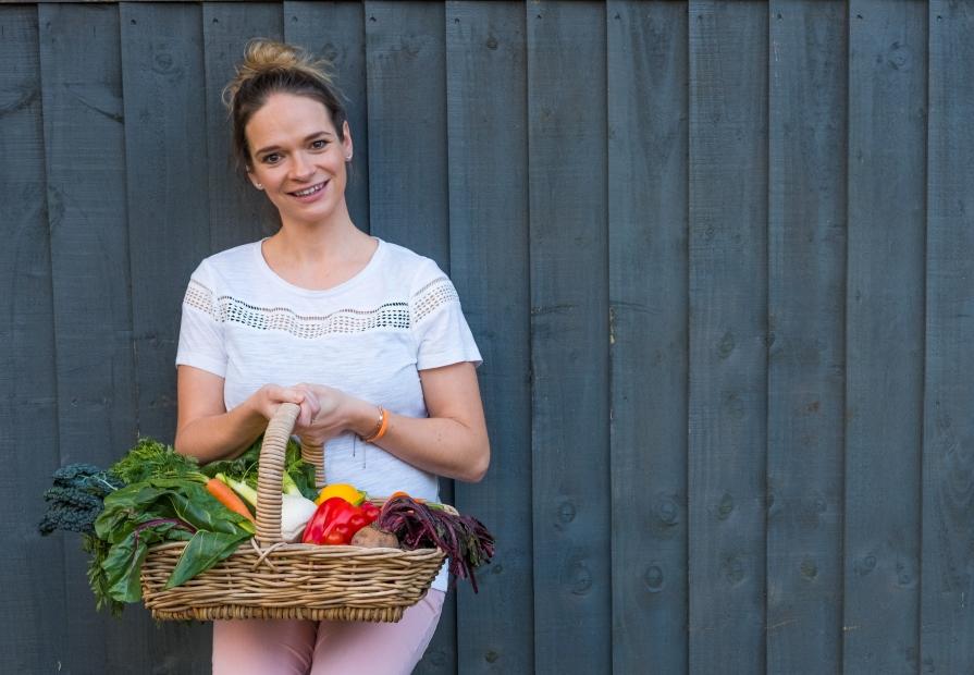 Lady in white t-shirt holding basket of vegetables, leaning against a great wall