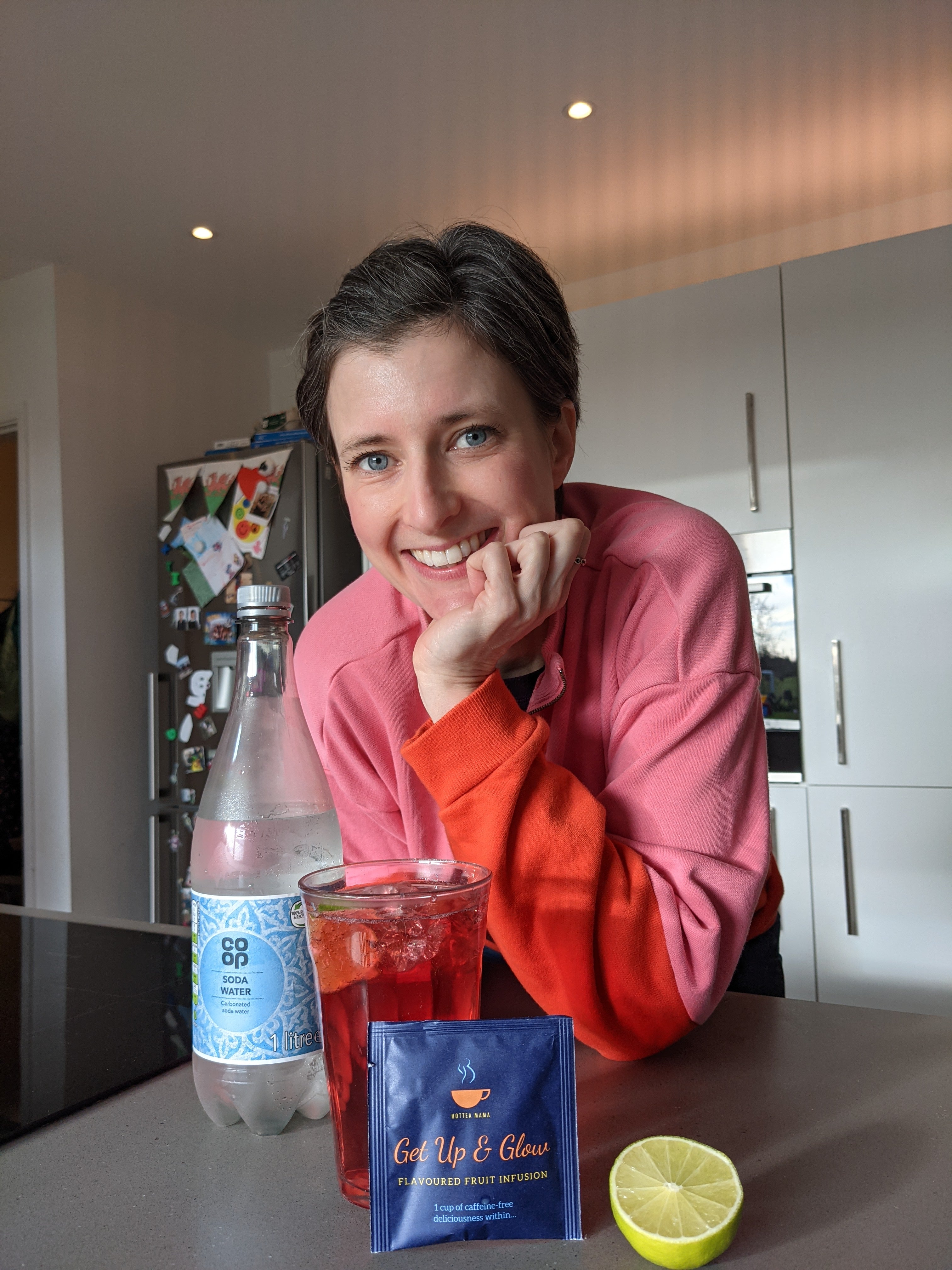 Woman leaning on kitchen side with get up and glow mocktail, soda, lime and tea bag on display.