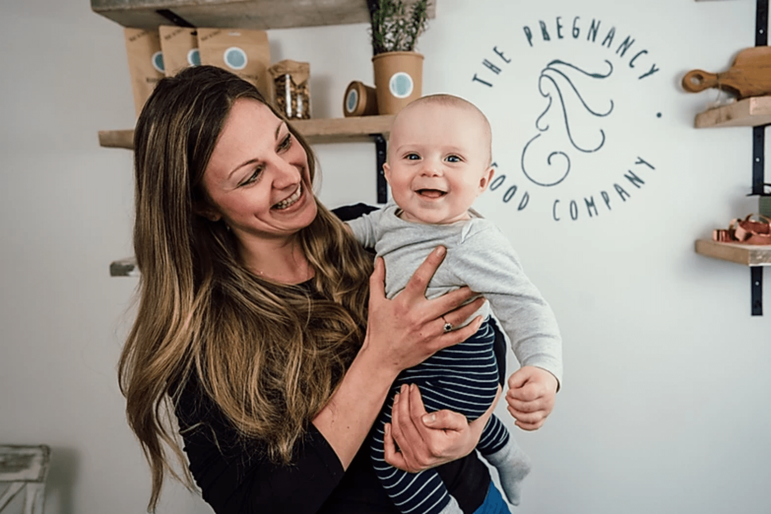 Lady holding a baby and smiling, in front of shelves with food on