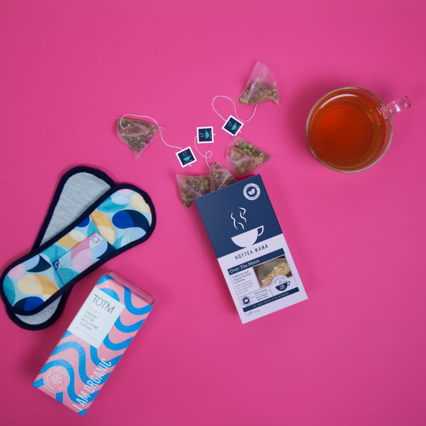 Endometriosis support tea box with tea bags coming out of the top, surrounded by period pads, tampons and a brewed cup of tea