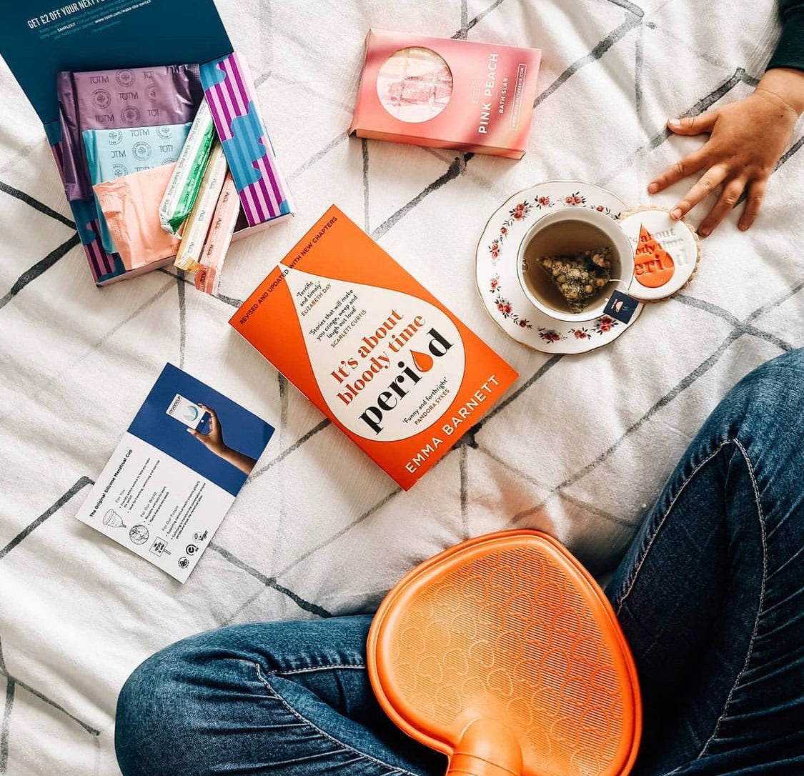 Lady's legs crossed on bed with hot water bottle, book about periods, a cup of Over the Moon menstruation tea and period periods.