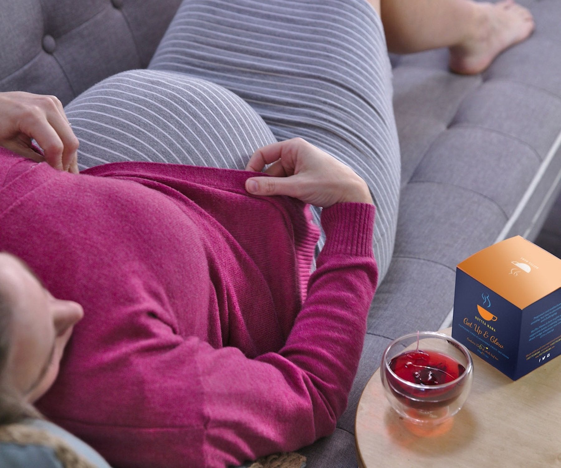 Pregnant lady in grey dress and pink cardigan, lying on sofa, with cup of Get Up & Glow tea on table next to her