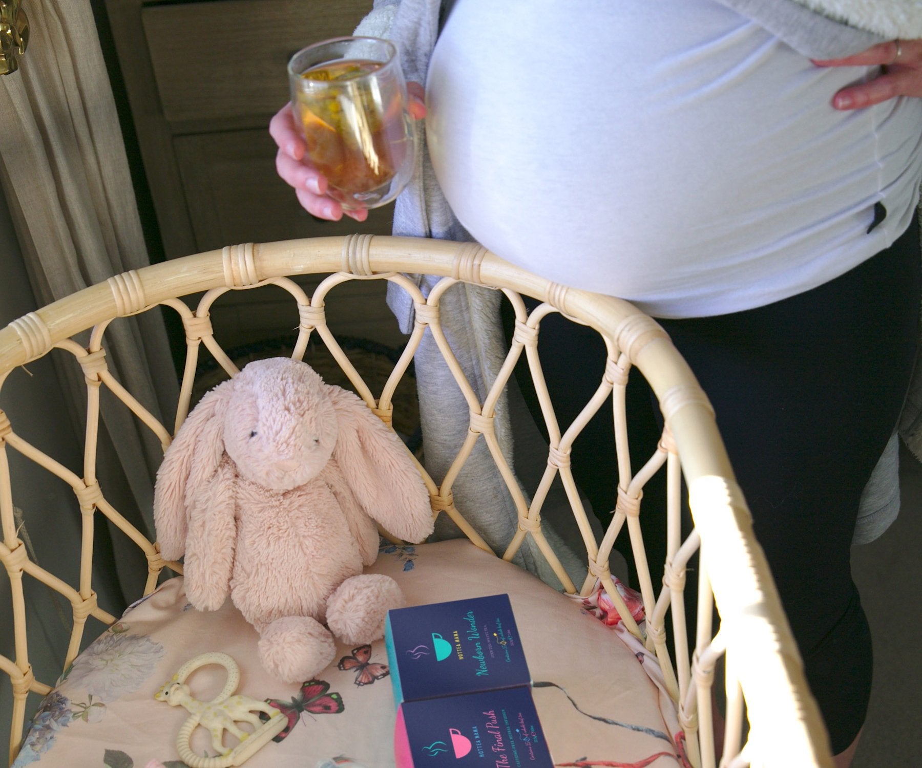Pregnant lady stood by cot, holding a cup of tea and looking at cuddly toys and tea packs inside the crib
