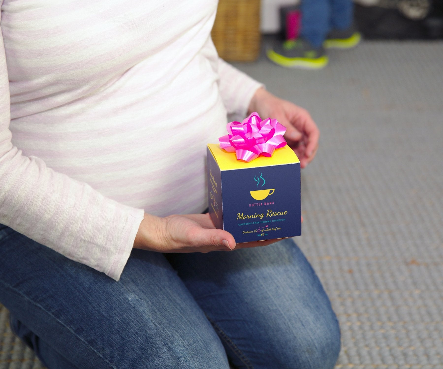 Pregnant lady knelt down, holding a pack of Morning Rescue tea with a pink bow on top