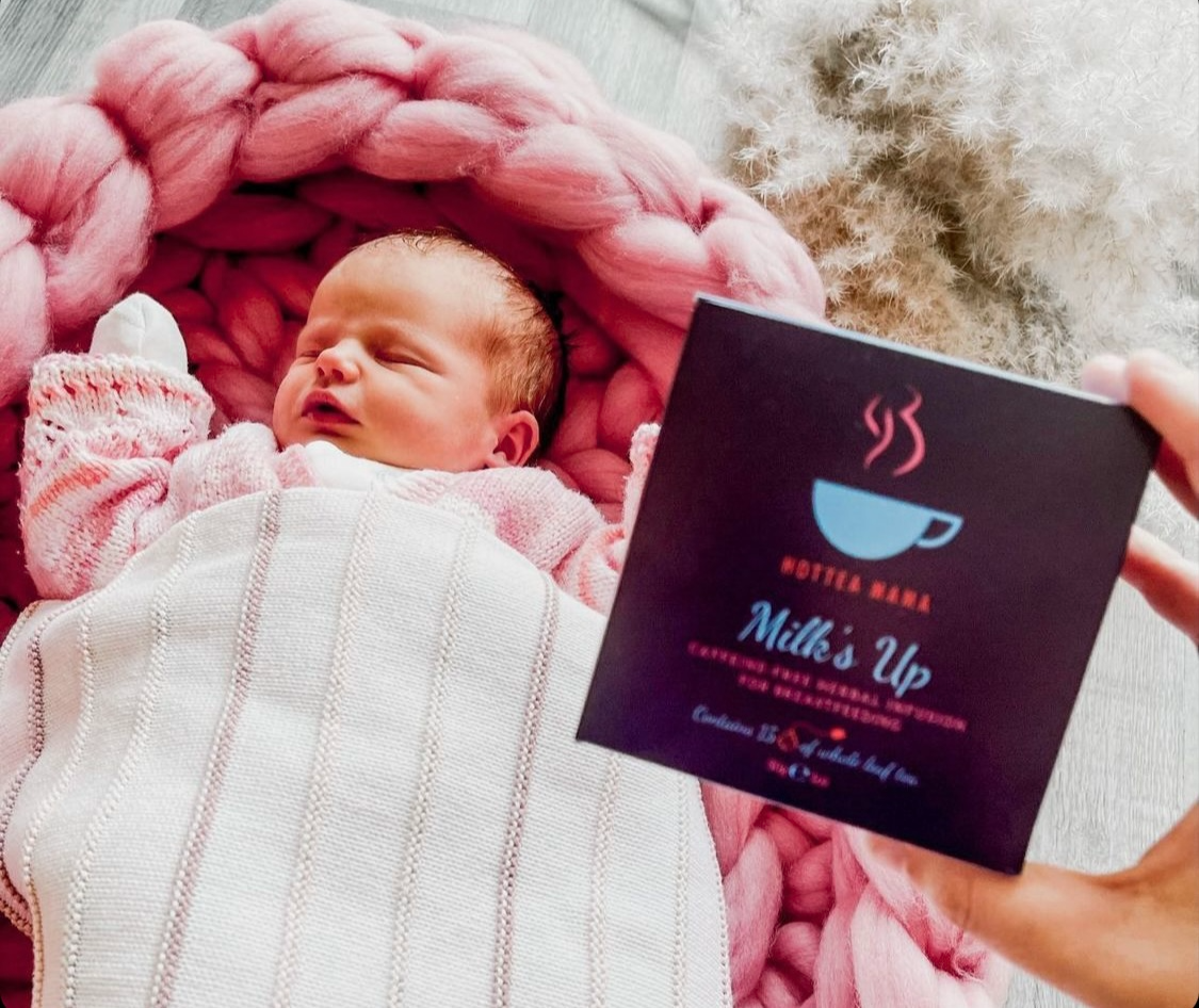 Baby in pink cot with a hand holding Milk's Up breastfeeding tea pack close to camera