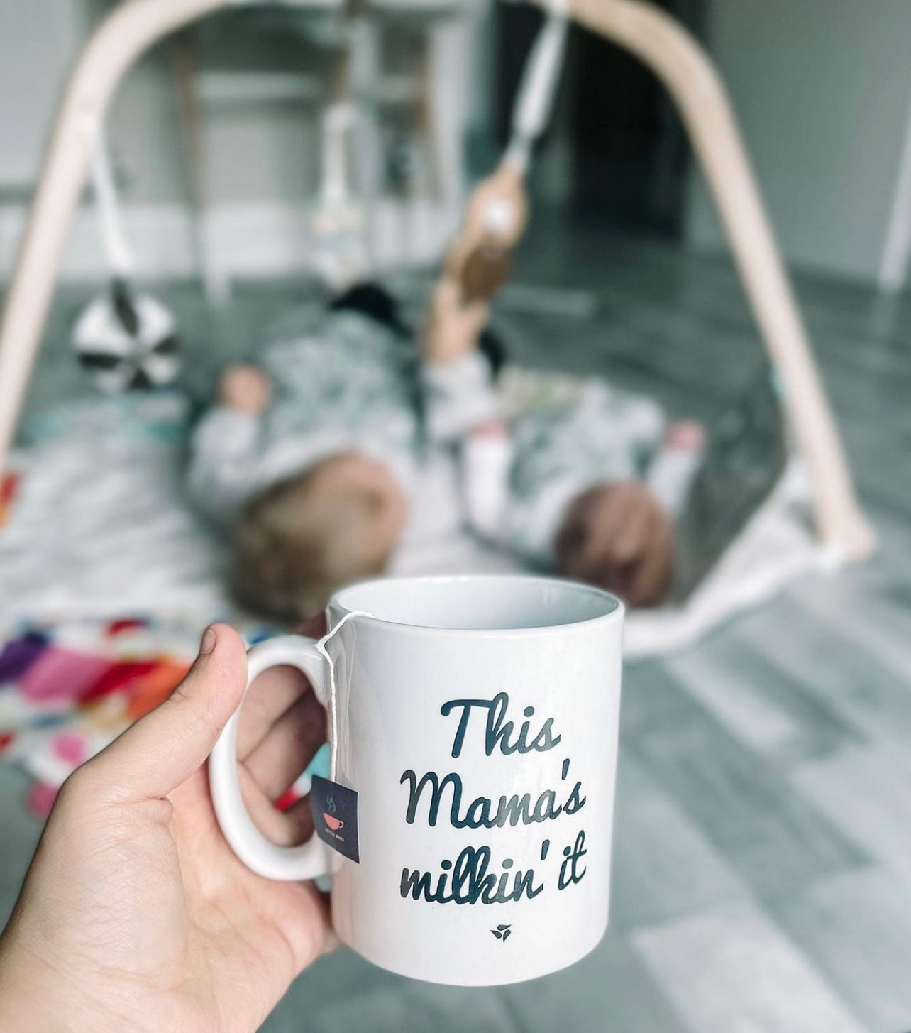 Babies on a play mat with mum watching while holding a cup of tea saying 'this mum's milkin' it' 