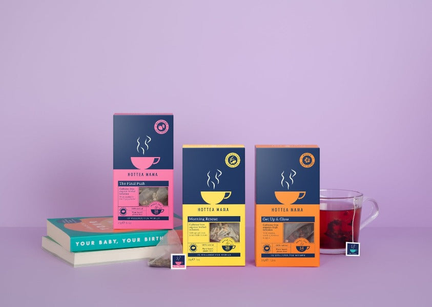 Three packs of pregnancy tea on a purple background, with The Final Push raspberry leaf tea sat of pregnancy books, Morning Rescue pack with tea bags next to it, and Get Up & Glow by a brewed cup of fruit tea
