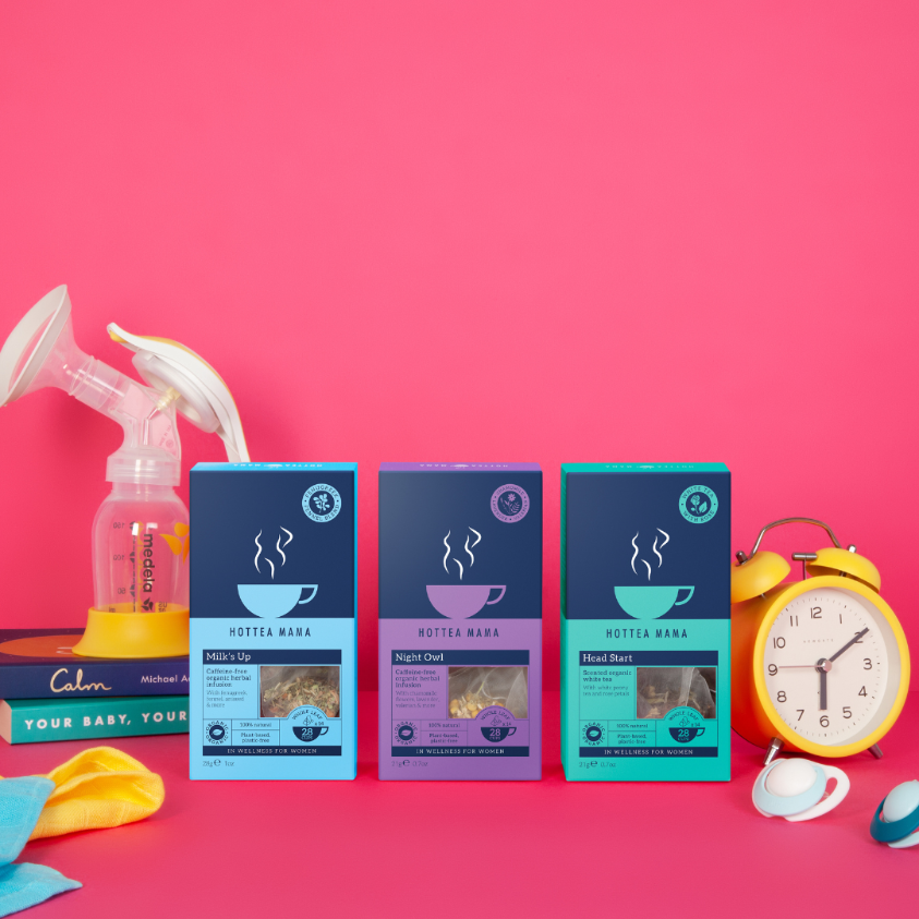 Three packs of HotTea Mama tea against a pink background, with a clock on one side and a breast pump and parenting books on the other.  The teas are a blue pack of Organic Milks Up breastfeeding tea, a purple pack of Organic Night Owl tea and a turquoise pack of Head Start white tea