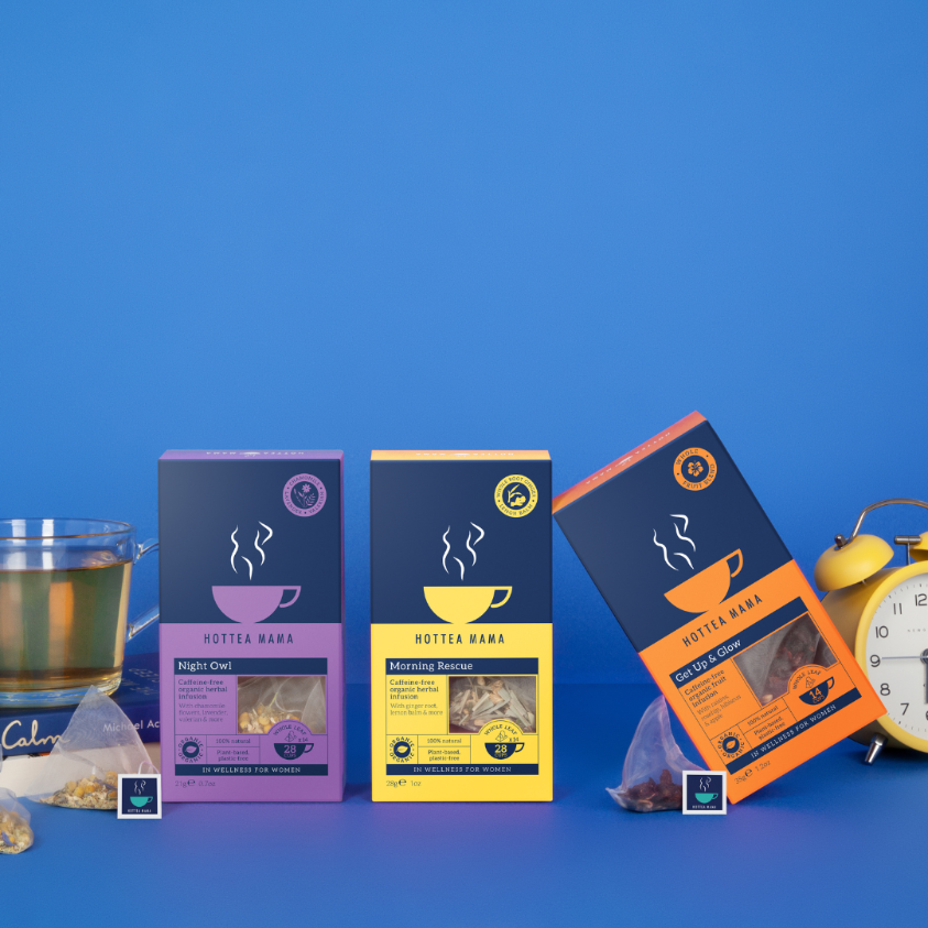 Three new mum support teas on a blue background - a brewed cup of organic Night Owl tea on top of a book about calm, Morning Rescue tea pack in middle of the shot and a pack of Get Up And Glow fruit tea on an angle next to an alarm clock