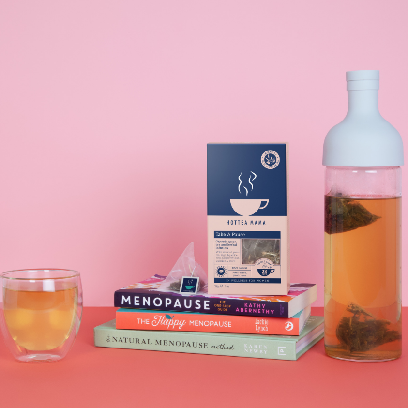 A pile of books on menopause with HotTea Mama's organic Take A Pause menopause tea say on top of them, with a cold brew bottle of iced tea and a cup of infused tea